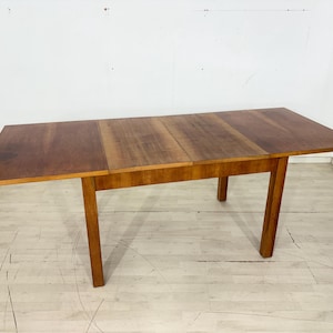 Può includere: Un tavolo da pranzo rettangolare in legno con una calda finitura marrone. Il tavolo presenta una superficie liscia e piana composta da tre sezioni distinte, che suggeriscono un design estensibile, sostenuto da quattro gambe in legno dritte e robuste.