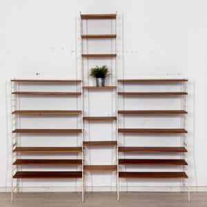Peut inclure: Un système d'étagères modulaire avec plusieurs sections. Les étagères sont en bois foncé et soutenues par une structure métallique blanche. Une petite plante en pot est posée sur l'une des étagères. L'ensemble est contre un mur blanc.