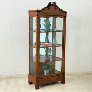 Peut inclure: Vitrine en bois vintage avec portes et étagères en verre. L'armoire est remplie d'objets décoratifs, notamment des plantes, des vases et une petite voiture jouet. Le meuble a un dessus décoratif et est en bois foncé.