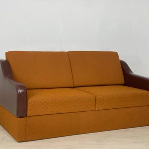 Może przedstawiać: Dwuosobowa brązowo-pomarańczowa sofa z brązowymi skórzanymi podłokietnikami. Sofa ma pikowaną tapicerkę.
