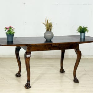 Peut inclure: Une table antique brun foncé avec un plateau rectangulaire et des pieds incurvés. La table a un tiroir et est décorée de petites plantes en pot et d'un vase. La table mesure environ 152 cm de long.