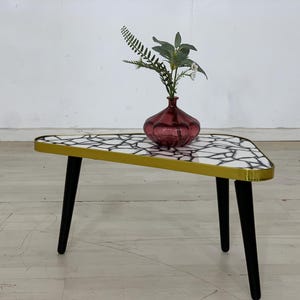 Peut inclure: Une table triangulaire avec un plateau à motifs blancs et noirs, une bordure dorée et des pieds noirs. Un vase en verre rose avec une plante verte feuillue est posé sur la table.