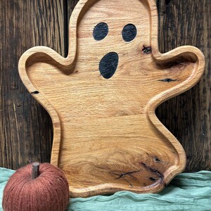 May include: A wooden ghost-shaped serving tray with black painted eyes and mouth. The tray is sitting on a green fabric surface next to a brown yarn ball.