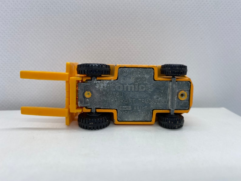 Pu&ograve; includere: Carrello elevatore a forche giocattolo giallo con ruote nere. La parte inferiore del camion ha il testo "Tomica 1/52 4  Komatsu Forklift Truck Made in Japan" stampato su di esso.