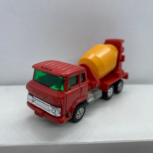May include: A red toy cement mixer truck with a yellow rotating drum. The truck has a green windscreen and black wheels.