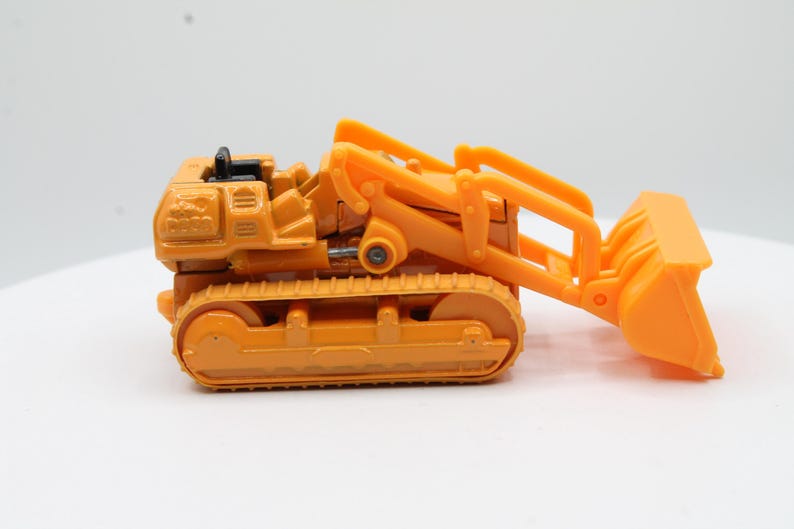 May include: Orange toy bulldozer with a black accent on top. The bulldozer has a front scoop and a track system for movement. The toy is made of plastic and is designed to resemble a construction vehicle.