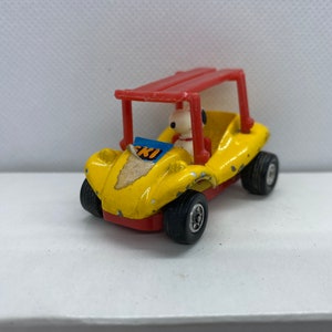 May include: A yellow toy dune buggy with a red roll cage and a white figure of Snoopy behind the wheel. The buggy has a blue and white sticker that says "SKI" on the front.
