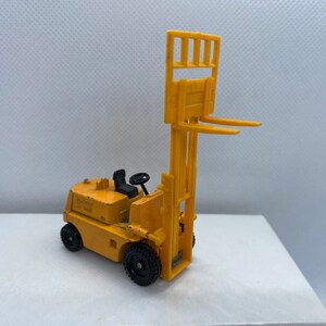 Pu&ograve; includere: Un carrello elevatore giocattolo giallo con ruote nere e un volante nero. Il carrello elevatore ha una piattaforma rialzata con una gabbia di metallo.