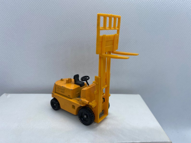 Pu&ograve; includere: Un carrello elevatore giocattolo giallo con ruote nere e un volante nero. Il carrello elevatore ha una piattaforma rialzata con una gabbia di metallo.