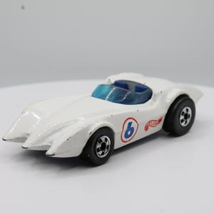 Può includere: Un'auto da corsa giocattolo bianca con parabrezza blu e ruote nere. L'auto ha il numero 6 in un cerchio blu con bordo rosso e il logo Hot Wheels. L'auto è su una superficie bianca.