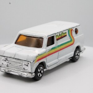 May include: A white toy van with a silver front grill and black wheels. The van has a rainbow stripe design on the side with the text "Rain Bow". The van is a vintage collectible toy.