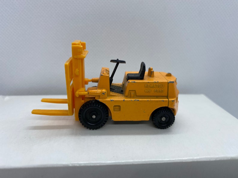 Pu&ograve; includere: Un carrello elevatore giocattolo giallo con ruote nere e un volante nero. Il carrello elevatore ha un sedile nero e dice "KOMATSU 650 5000" sul lato.