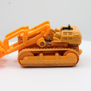 May include: Orange toy bulldozer with a front bucket and caterpillar tracks. The construction vehicle is made of plastic and sits on a white surface. The bulldozer's blade is lowered, ready for action.
