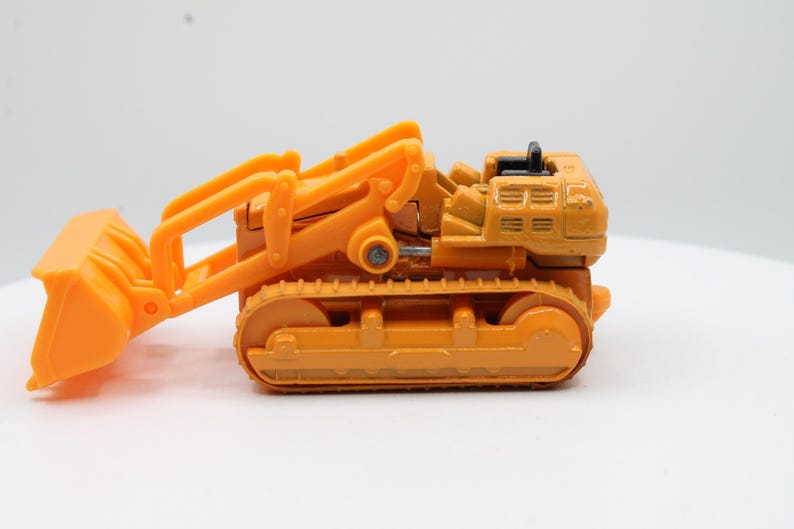 May include: Orange toy bulldozer with a front bucket and caterpillar tracks. The construction vehicle is made of plastic and sits on a white surface. The bulldozer's blade is lowered, ready for action.