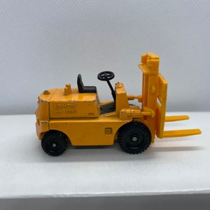Pu&ograve; includere: Un carrello elevatore giocattolo giallo con ruote nere e un volante nero. Il carrello elevatore ha la scritta "KOMATSU  FG10" sul lato.