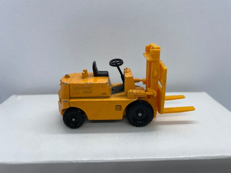 Pu&ograve; includere: Un carrello elevatore giocattolo giallo con ruote nere e un volante nero. Il carrello elevatore ha la scritta "KOMATSU  FG10" sul lato.