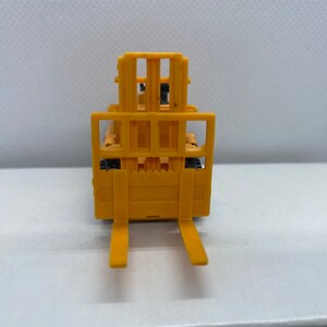 Pu&ograve; includere: Un carrello elevatore giocattolo giallo con una forca sollevata e una griglia nera. Il carrello elevatore &egrave; su una superficie bianca.