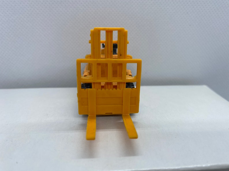 Pu&ograve; includere: Un carrello elevatore giocattolo giallo con una forca sollevata e una griglia nera. Il carrello elevatore &egrave; su una superficie bianca.