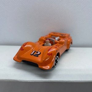 May include: Orange toy racing car with the number 12 on the side and a small figure of a driver inside. The car has a red and orange flame design.