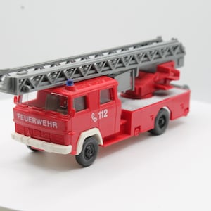 Op de afbeelding: Een rode speelgoed brandweerwagen met een grijze ladder. De vrachtwagen heeft zwarte wielen, witte accenten en het woord "FEUERWEHR" in het wit. Het nummer "112" is ook zichtbaar.
