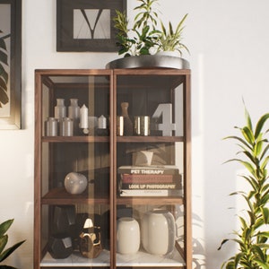K&ouml;nnte beinhalten: Ein Holzvitrine mit Glasb&ouml;den, die verschiedene Dekorationsartikel zeigt, darunter Kerzen, Vasen, B&uuml;cher und eine Keramik-Kugel. Die Vitrine steht vor einer wei&szlig;en Wand mit einem gerahmten Druck und einer Topfpflanze.