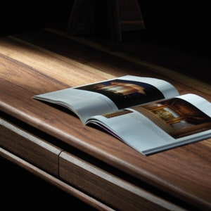 Puede incluir: Primer plano de un escritorio de madera con una revista abierta encima. El escritorio tiene un acabado de madera natural y un cajón en el lateral. La revista está abierta en una página con una imagen de un edificio.