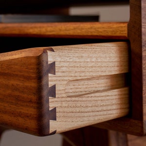 May include: Close-up of a wooden drawer with dovetail joints. The drawer is made of light-colored wood and is set within a darker wood frame. The image includes the text "D&Iacute;AZ MARENTES STUDIO The Art of the Functional".