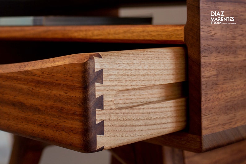 May include: Close-up of a wooden drawer with dovetail joints. The drawer is made of light-colored wood and is set within a darker wood frame. The image includes the text "D&Iacute;AZ MARENTES STUDIO The Art of the Functional".