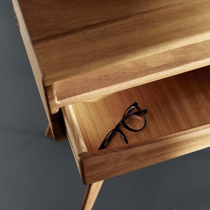 May include: A wooden nightstand with a drawer slightly open, revealing a pair of black-framed eyeglasses. The nightstand has a natural wood finish and a minimalist design. The legs are angled, and the drawer has a simple handle. The overall aesthetic is modern and functional.