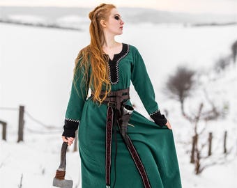 Op de afbeelding: Een vrouw in een groene jurk met een zwarte riem en een rode en witte bies staat in een besneeuwd veld en houdt een bijl vast. Ze heeft lang rood haar en kijkt naar rechts.