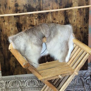 Puede incluir: Una silla plegable de madera con una piel de pelo de color claro sobre ella. La silla tiene un acabado de madera natural y un diseño de patas cruzadas. La piel es gris y blanca. Un objeto azul claro está unido a la piel.