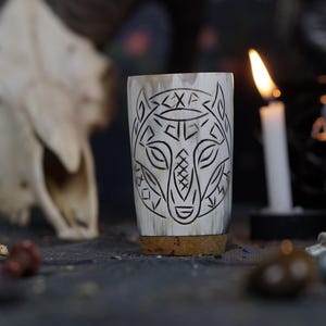 Può includere: Un corno da bere bianco con un disegno di lupo nero inciso sul lato. Il corno è appoggiato su una superficie scura con un teschio e altri oggetti sullo sfondo.