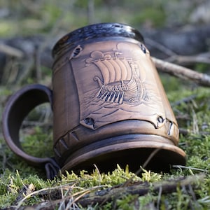 Könnte beinhalten: Eine braune Keramiktasse mit einem Reliefdesign eines Wikingerschiffs mit Segeln. Die Tasse hat einen Henkel und steht auf einem Bett aus grünem Moos.