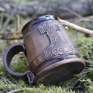 Peut inclure: Une tasse en céramique brune avec un motif en relief d'un marteau et d'un nœud. La tasse est recouverte d'un glaçage brun foncé et décorée d'un motif en relief d'un marteau et d'un nœud. La tasse est posée sur le côté sur un lit de mousse.