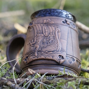 Könnte beinhalten: Eine braune Keramiktasse mit einem Reliefdesign eines Wikingerschiffs und seiner Besatzung. Die Tasse hat eine dunkelbraune Glasur und einen Henkel.
