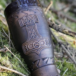 Può includere: Un corno potorio color bronzo con un rilievo dettagliato del martello di Thor, Mjolnir. Il corno è cilindrico con una parte superiore svasata e una base decorativa. Lo sfondo mostra muschio e ramoscelli.