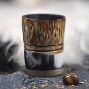 Può includere: Una tazza di legno fatta a mano con un design unico. La sezione superiore presenta linee verticali scolpite, mentre la sezione centrale presenta un motivo in bianco e nero. La tazza è marrone, nera e bianca, con un interno scuro.