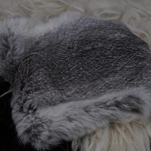 May include: A gray and white fur hat, likely made of rabbit fur, is displayed. The hat is a mix of gray and white, with a soft, textured appearance. The hat is resting on a white and black surface.