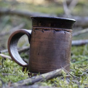 Könnte beinhalten: Handgefertigte braune Keramiktasse mit dunkelbraunem Rand und Henkel. Die Tasse hat eine strukturierte Oberfläche und steht vor einem Hintergrund aus Moos und abgefallenen Ästen. Diese rustikale Tasse ist perfekt für Heißgetränke.