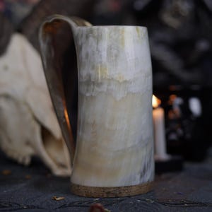 Può includere: Un boccale in corno fatto a mano con una tavolozza di colori naturale bianco sporco e beige. Il boccale presenta un manico grande e curvo e una base strutturata marrone chiaro. Lo sfondo include un teschio e altri elementi decorativi.