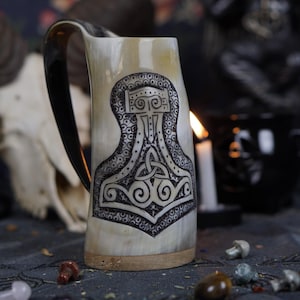 Könnte beinhalten: Ein weißer Trinkhornbecher mit einem schwarz-weißen Design aus einem Hammer und einem keltischen Knoten. Der Becher hat einen schwarzen Henkel.