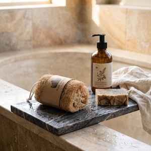 May include: A cylindrical loofah with a tan paper label reading Blue Ghost Acres, a rectangular bar of mottled soap, and an amber glass pump bottle with a white label featuring botanical illustrations, all resting on a dark grey marble tray.