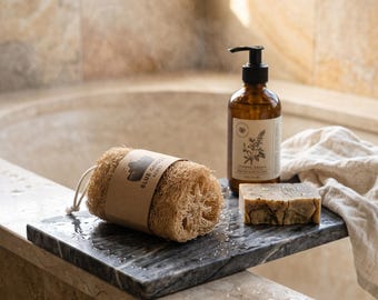 May include: A cylindrical loofah with a tan paper label reading Blue Ghost Acres, a rectangular bar of mottled soap, and an amber glass pump bottle with a white label featuring botanical illustrations, all resting on a dark grey marble tray.