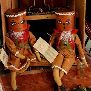 May include: Two handmade gingerbread people dolls with painted faces and white trim. The dolls are wearing red and green clothing and are sitting on a wooden shelf. The doll on the left is holding a sign that says "Hello My Name Is Gertie".