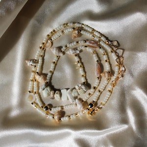 May include: A layered necklace with small white beads, gold-coloured accents, and seashells. The necklace has a central section with irregular, light-coloured stones. The clasp is gold-coloured. The jewellery is displayed on a satin surface.