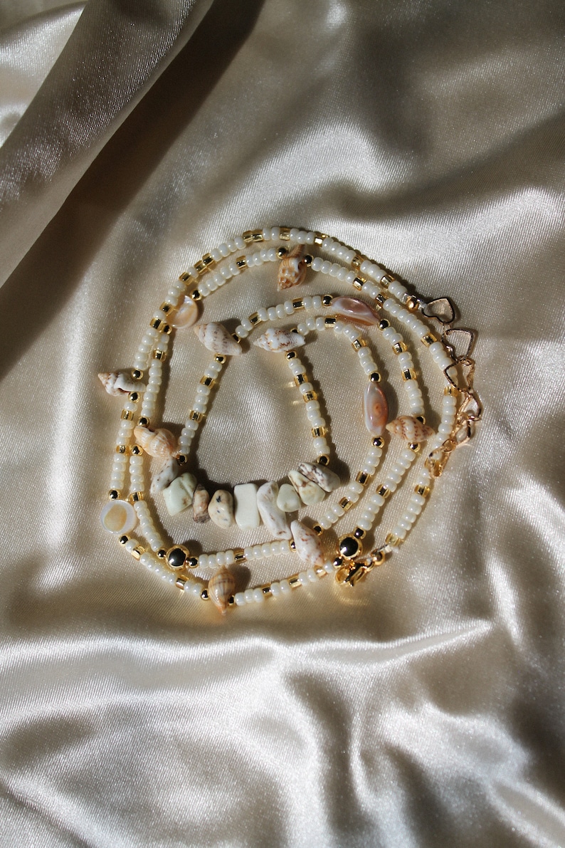 May include: A layered necklace with small white beads, gold-coloured accents, and seashells. The necklace has a central section with irregular, light-coloured stones. The clasp is gold-coloured. The jewellery is displayed on a satin surface.