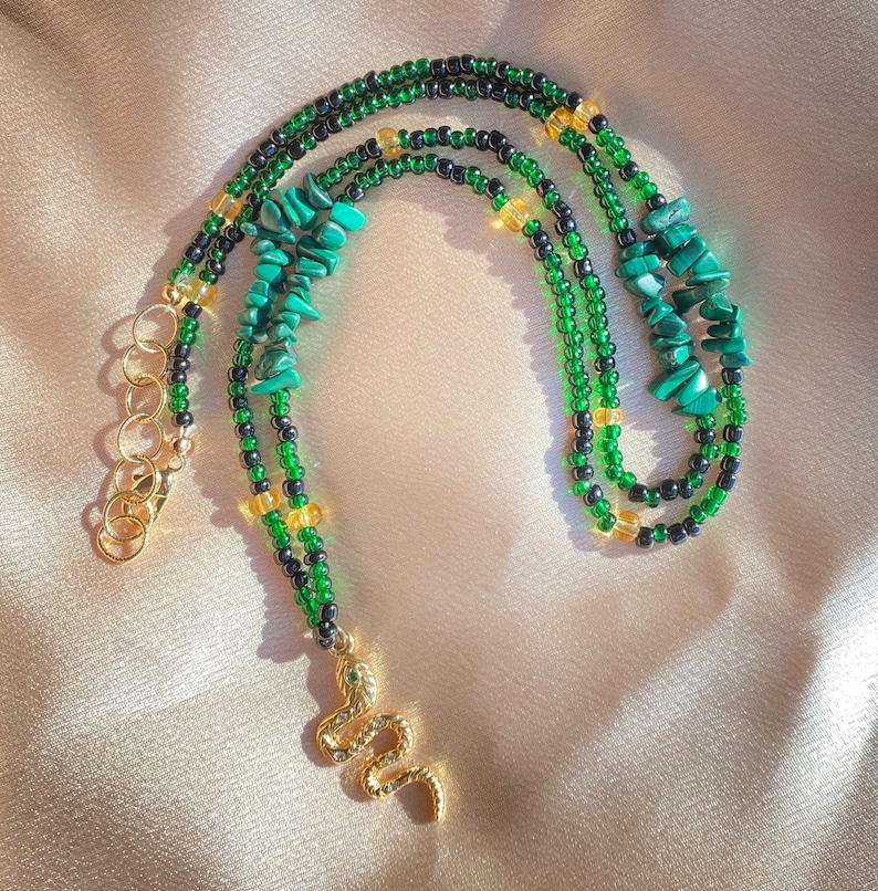 K&ouml;nnte beinhalten: Eine gr&uuml;ne und schwarze Perlenkette mit einem goldenen Schlangenanh&auml;nger. Die Kette ist mit gr&uuml;nen Malachit-Steinen und goldenen Perlen besetzt.