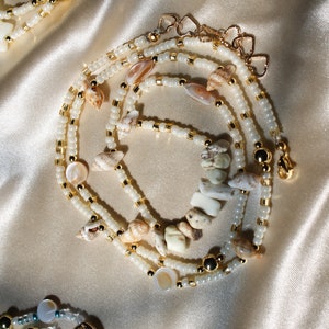 Op de afbeelding: Een gelaagde ketting met witte kralen, gouden accenten en natuurlijke schelpen. De ketting heeft een gouden sluiting en wordt gepresenteerd op een satijnen oppervlak. Het ontwerp bevat verschillende schelpvormen en -maten.