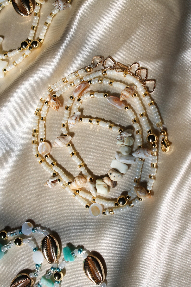 May include: A layered necklace with white beads, gold details, and seashells. The necklace has a gold clasp and is shown on a satin surface. The design incorporates various shell shapes and sizes.