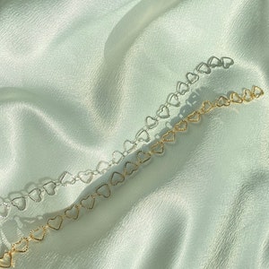May include: Two delicate heart-shaped chain necklaces, one silver and one gold, lay on a soft, white fabric background.